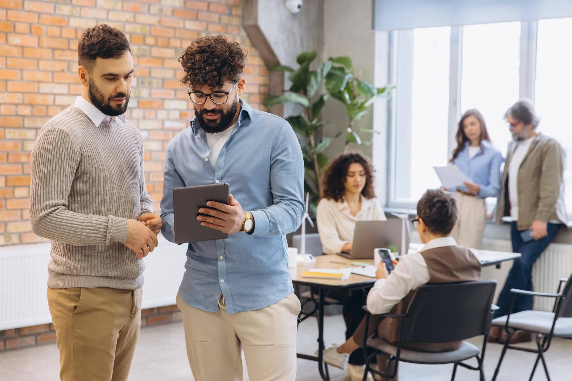 Two colleagues standing with a tablet in an open-plan office while others work at desks behind them